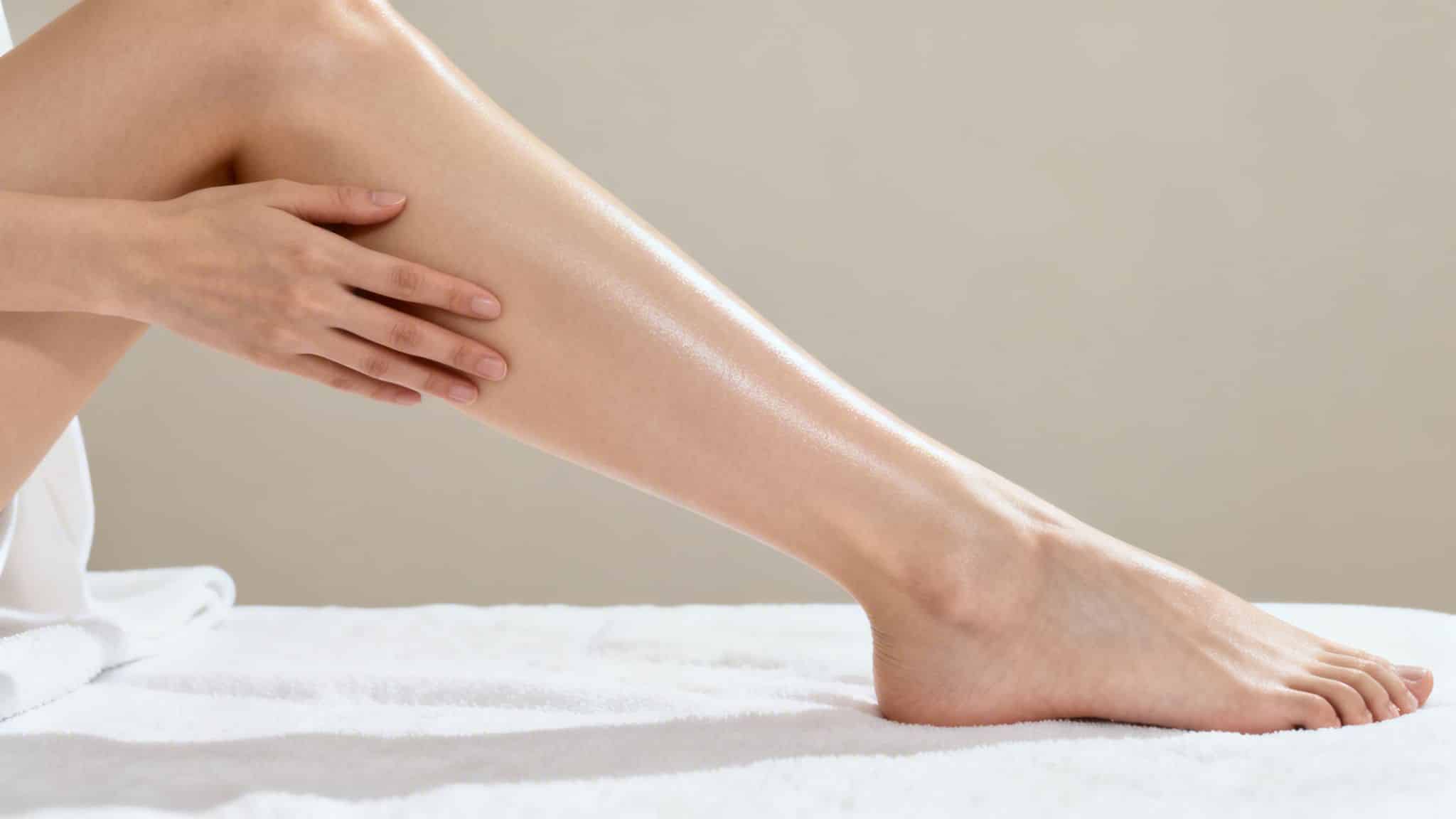 Main de femme appliquant de la lotion sur une jambe hydratée et brillante, posée sur une serviette blanche.