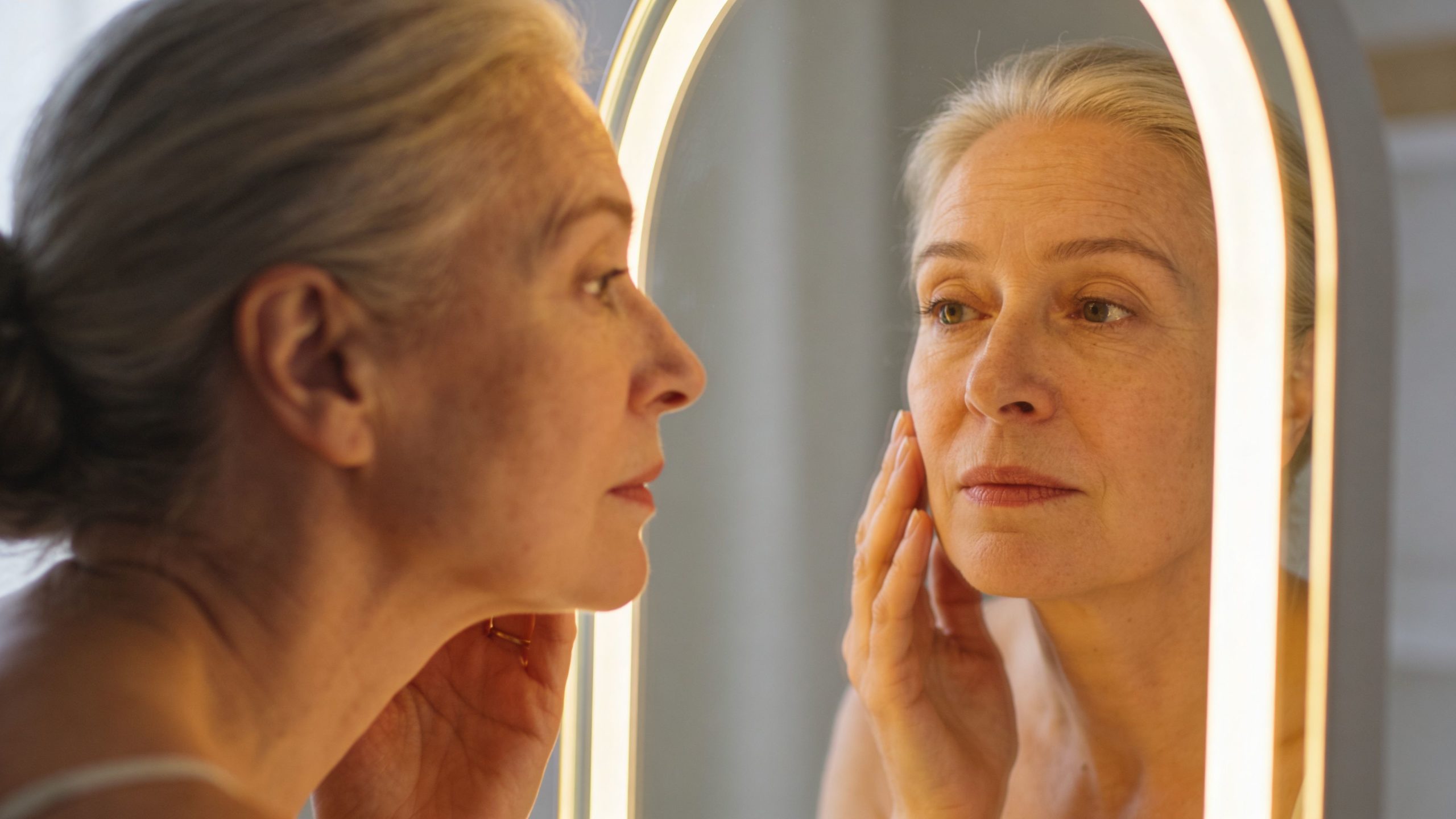 Une femme mûre aux cheveux gris se regarde attentivement dans un miroir lumineux après un peeling au phénol.