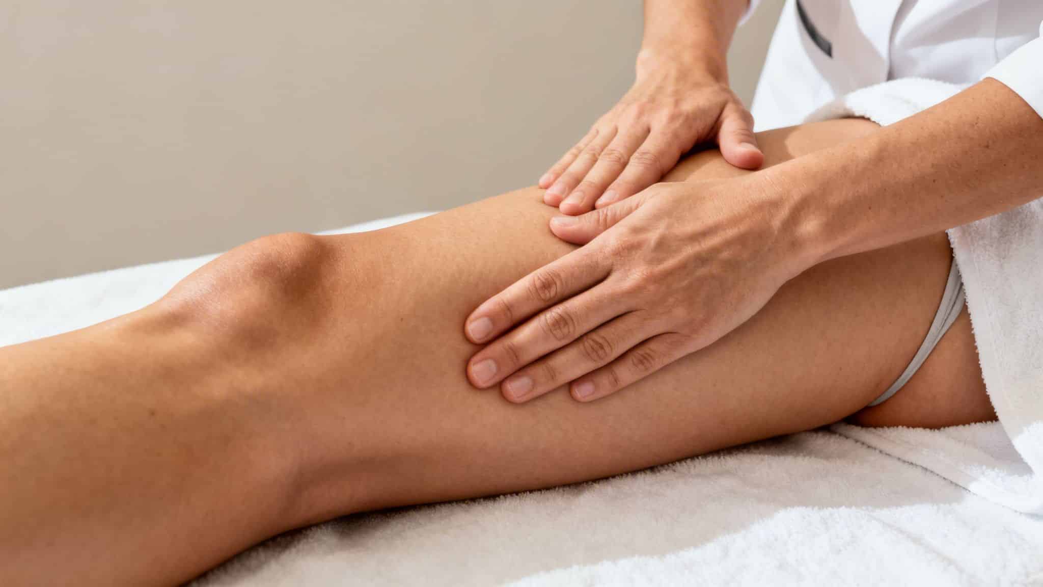 Deux mains massent délicatement la jambe d'une personne allongée sur une serviette blanche, vue de près.