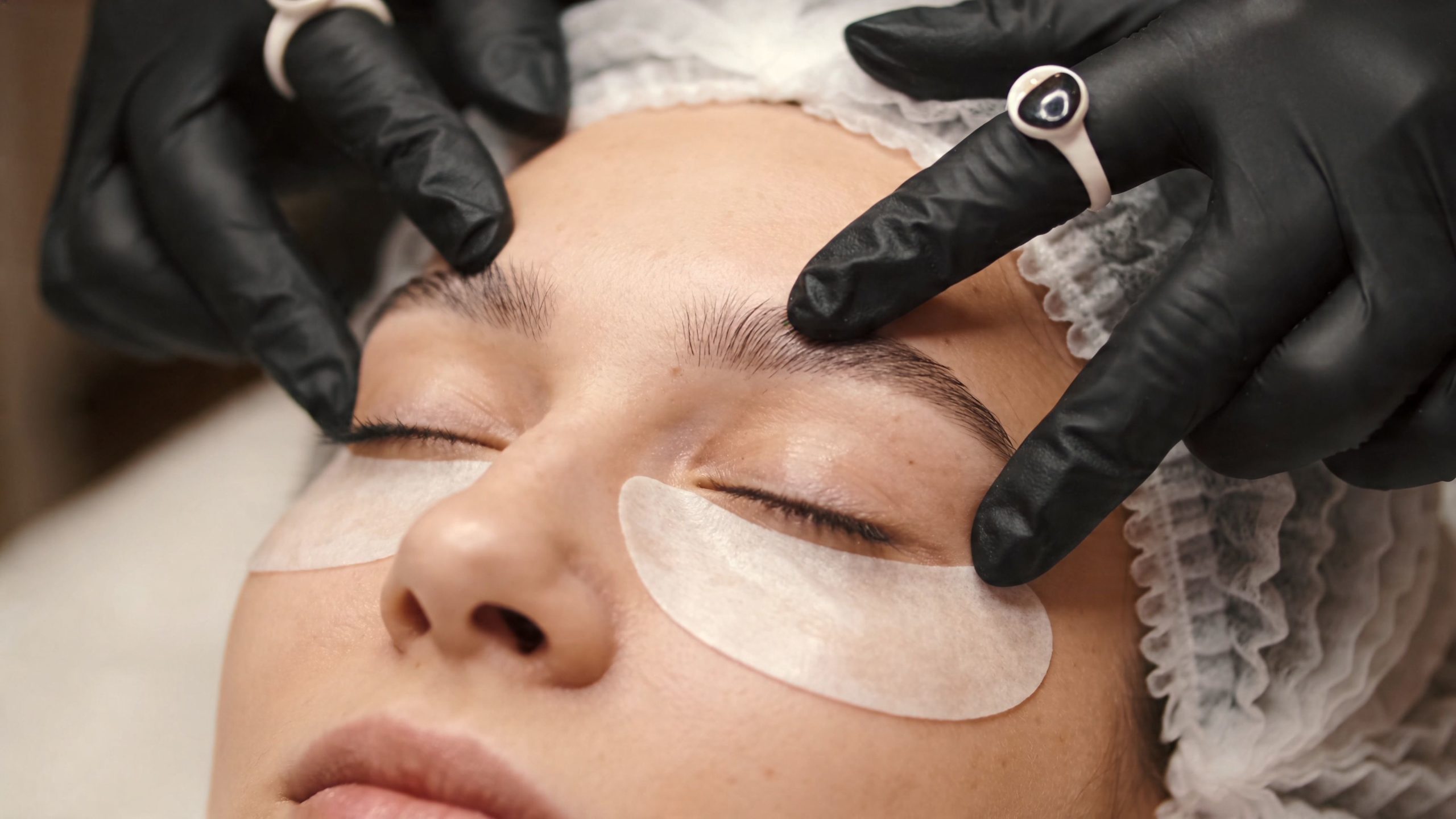 Une esthéticienne effectuant un soin des sourcils sur le visage d'une cliente portant des patchs anti-cernes.