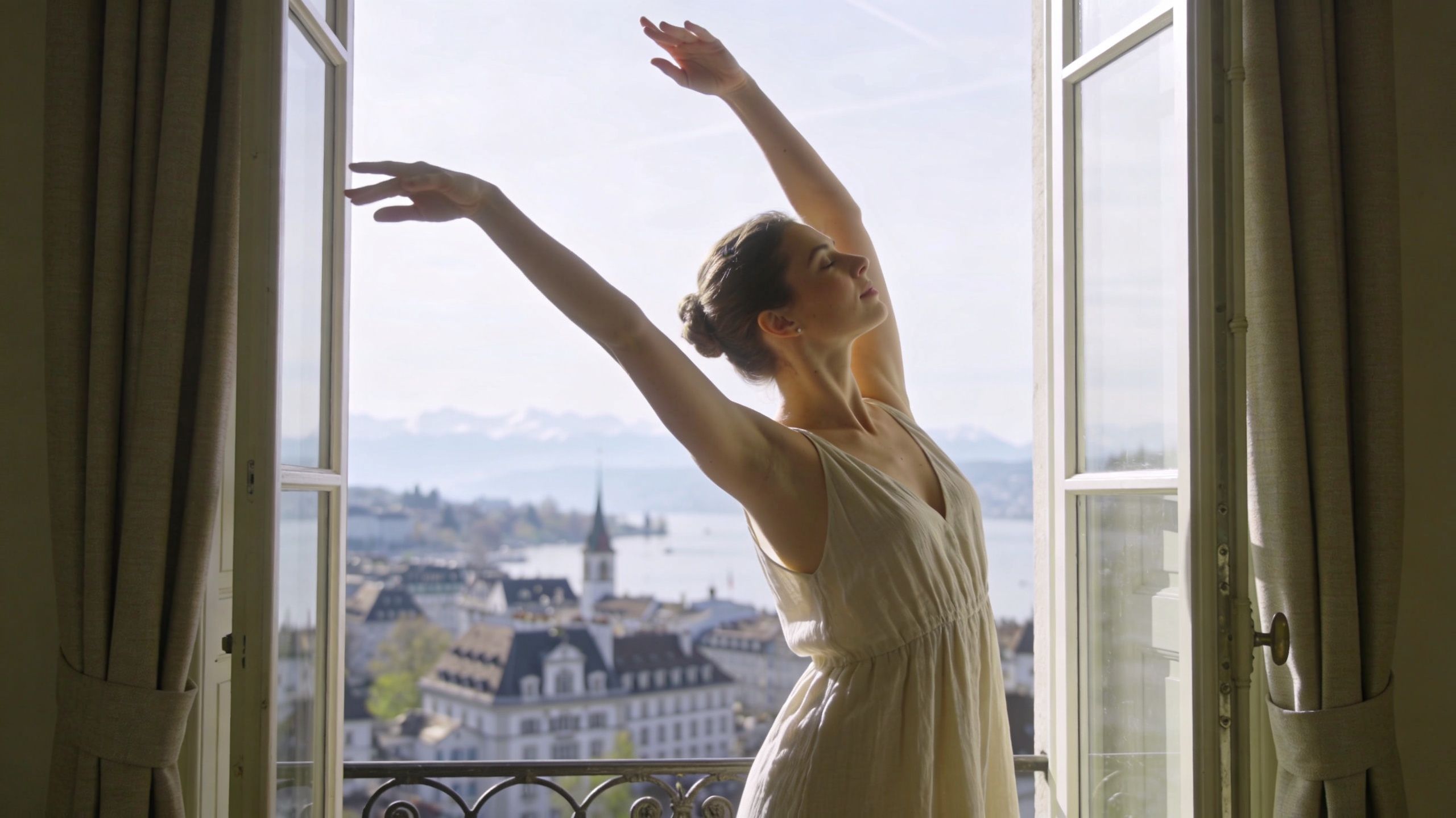 Une femme élégante en robe claire étirant ses bras devant une fenêtre surplombant le lac de Genève