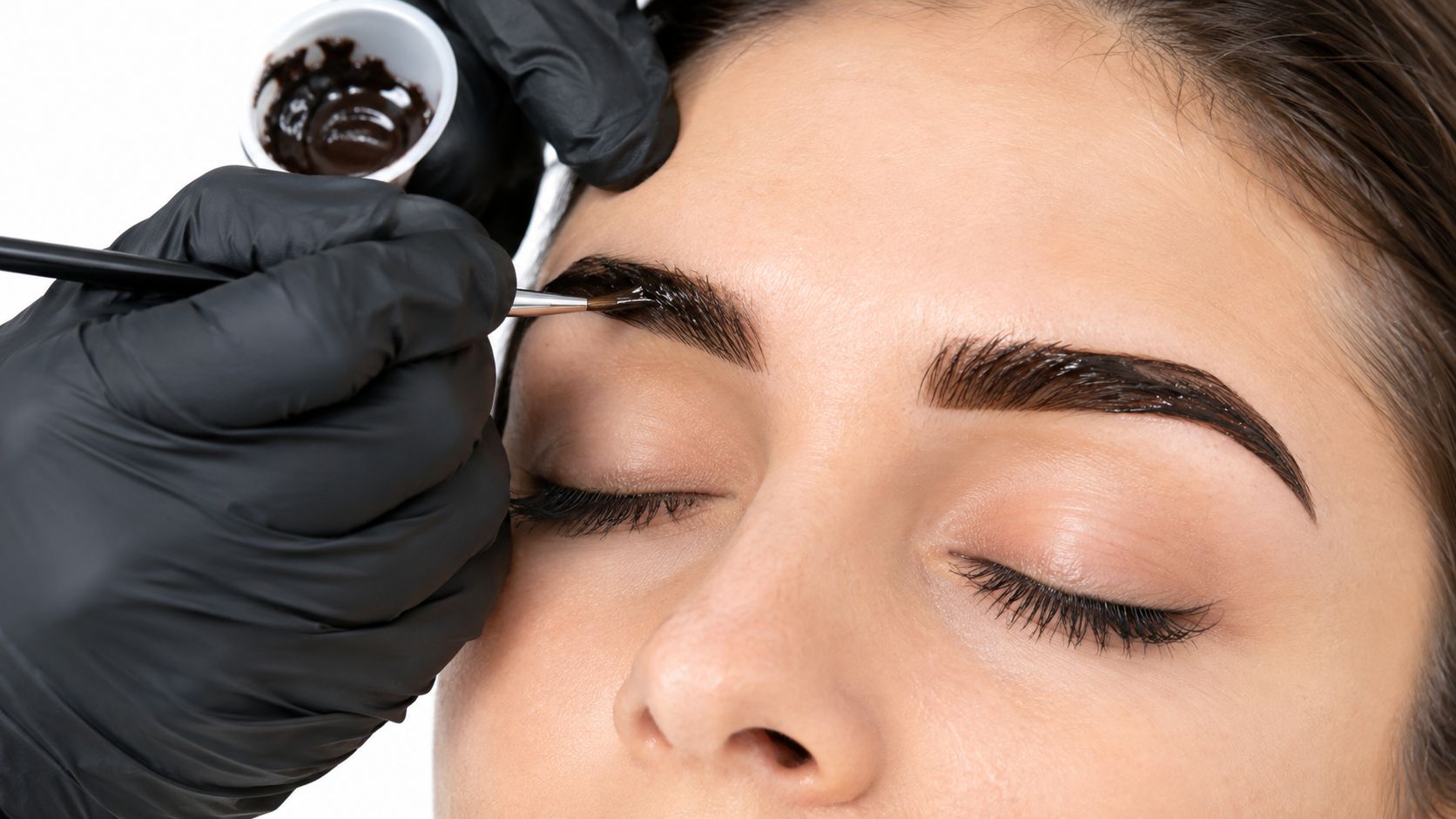 Une esthéticienne applique soigneusement de la teinture foncée sur les sourcils d'une cliente avec un pinceau fin.